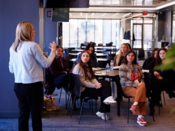 Media representative answering student's questions, with the students sitting at circular tables in a glass room.